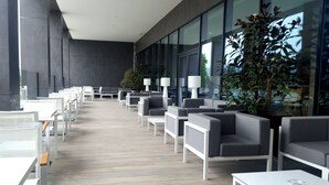 A modern outdoor seating area with grey sofas and white tables, large windows, and potted plants.