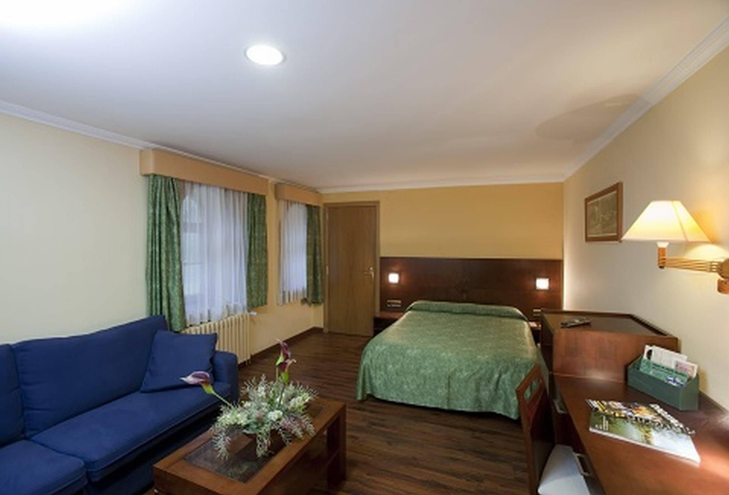 Habitación de hotel con sofá, cama con manta verde, mesita de noche de madera con lámpara y ventana con cortinas.