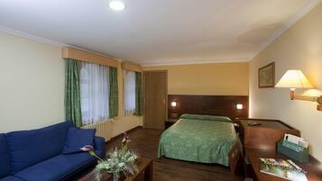 Habitación de hotel con sofá, cama con manta verde, mesita de noche de madera con lámpara y ventana con cortinas.
