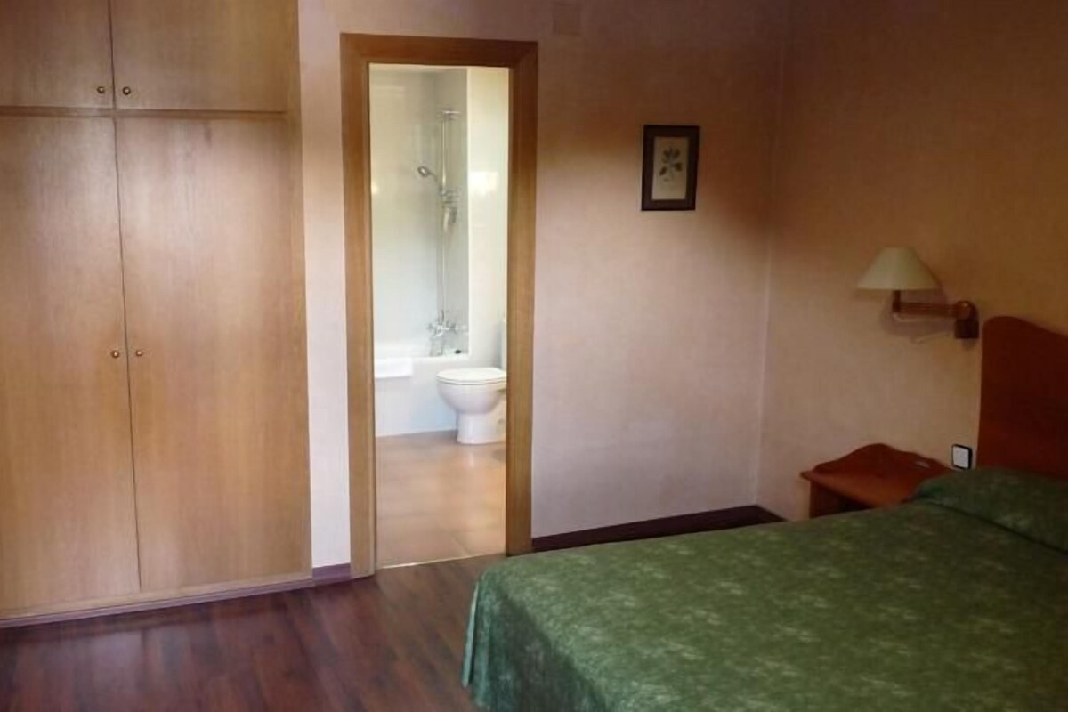 A bedroom with a wooden wardrobe, a green bed, a wooden chair, a bedside table, and a bathroom visible through an open door.