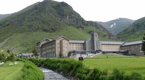 Un edificio grande con una torre destacada, probablemente un hotel, frente a un paisaje montañoso y con un río en primer plano.