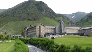 Un edificio grande con una torre destacada, probablemente un hotel, frente a un paisaje montañoso y con un río en primer plano.