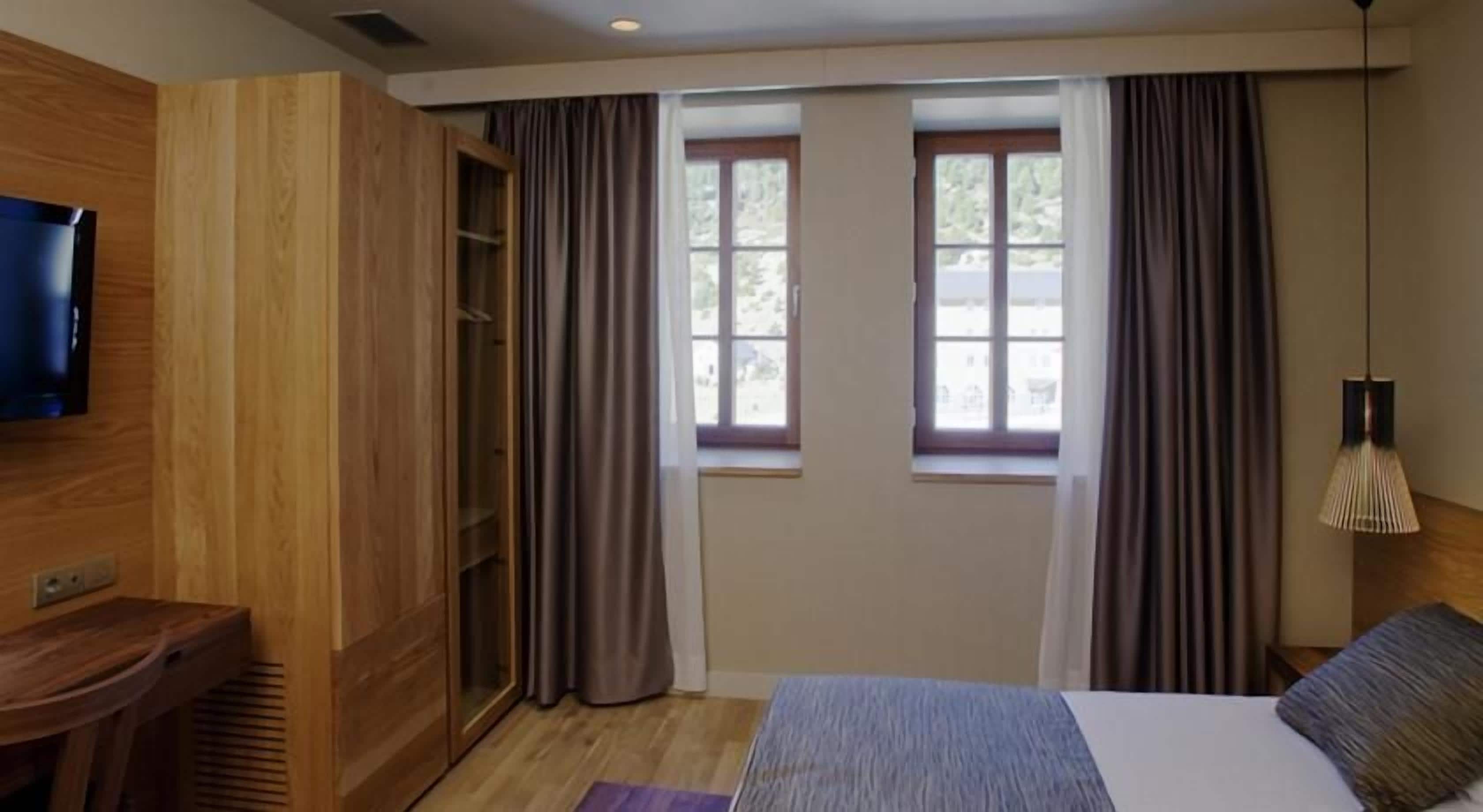 Habitación de hotel con una cama, televisión, armario de madera, ventana con cortinas y una lámpara.