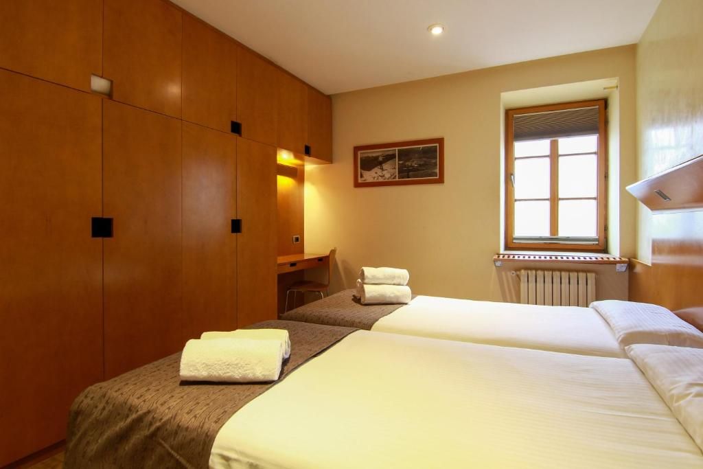 A hotel room with a large wardrobe, a desk, a window with a view, and two beds.
