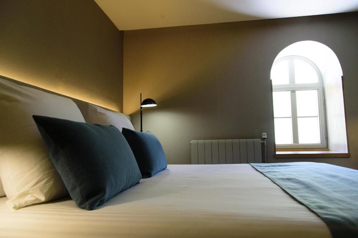 A bedroom with a bed, pillows, a lamp, a radiator, and a window with an arch.