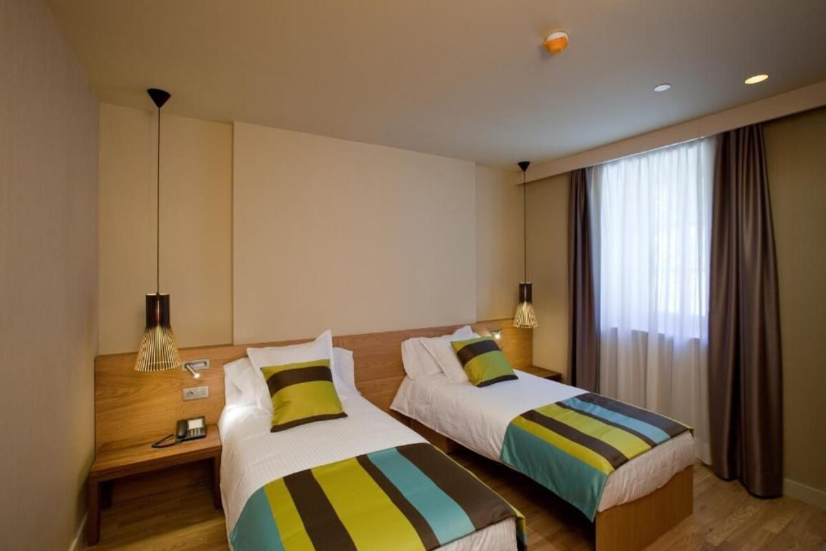 A hotel room with two single beds, wooden headboards, bedside tables with lamps, and a large window with curtains.