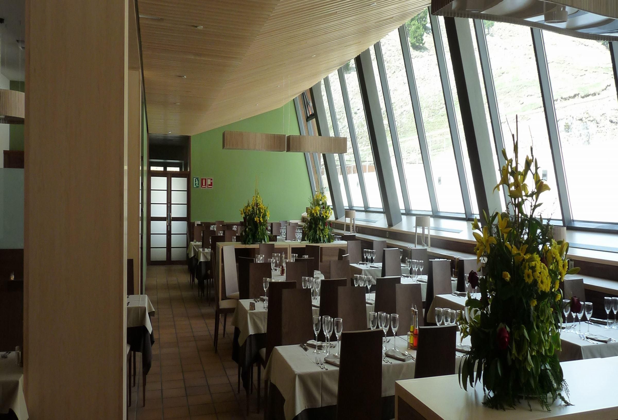 Un restaurante con varias mesas, sillas y amplios ventanales que dejan entrar luz natural.