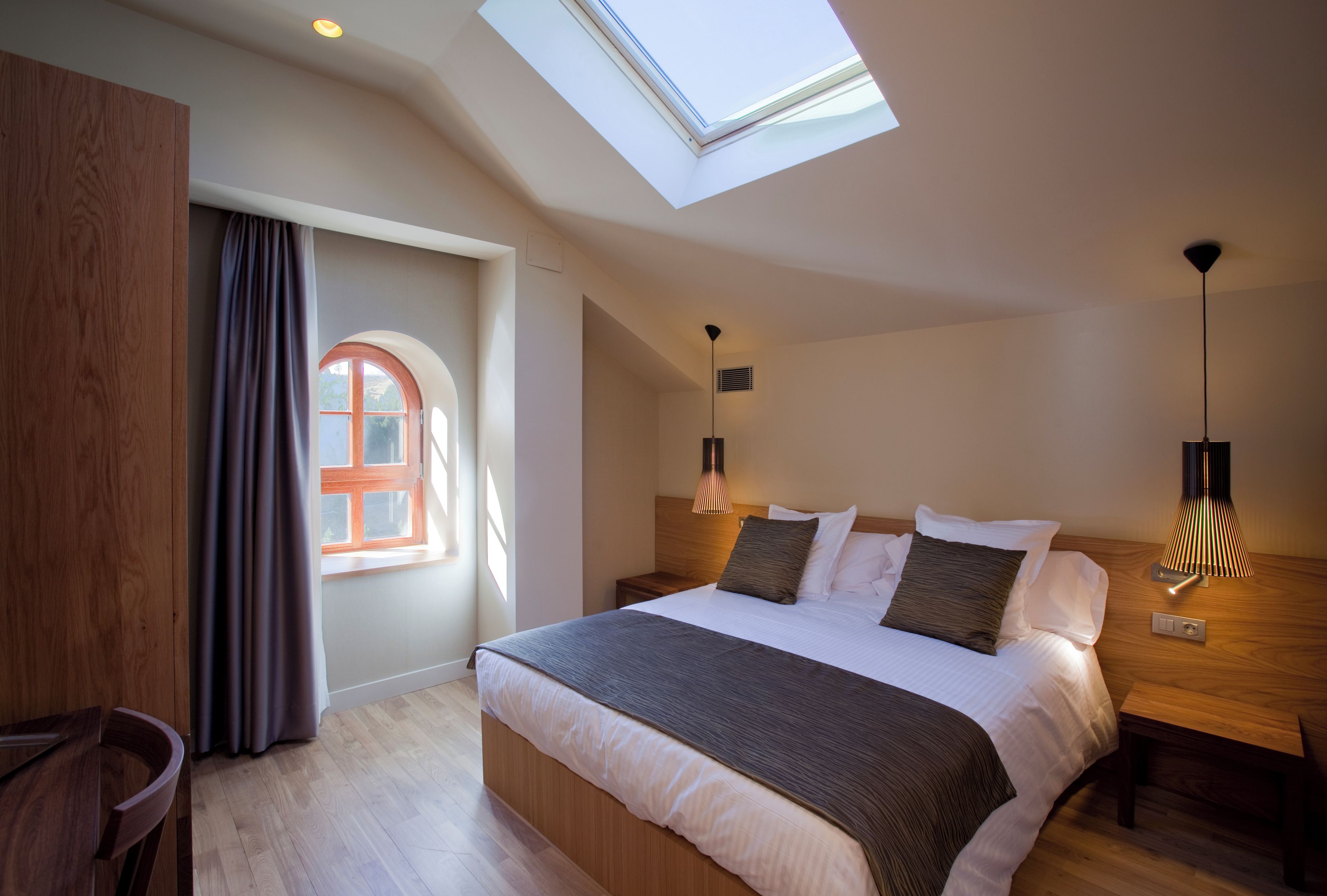 Un dormitorio moderno con tragaluz, una cama con muchos almohadones, lámparas de noche y vistas a través de una ventana.