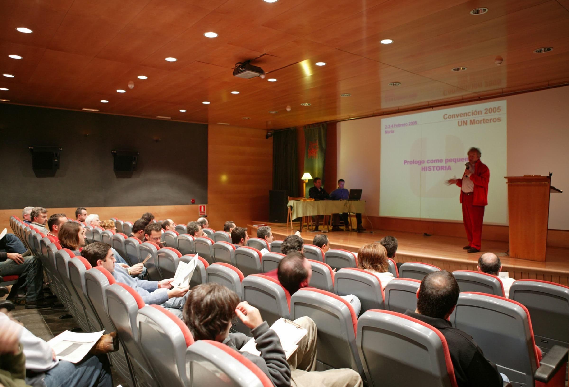 Un salón de conferencias con filas de sillas mirando hacia un escenario donde un orador está presentando.