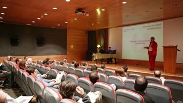 Un salón de conferencias con filas de sillas mirando hacia un escenario donde un orador está presentando.