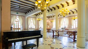 Un piano de cola imponente en un vestíbulo amplio con suelo de baldosas, ventanas grandes con cortinas y múltiples candelabros.