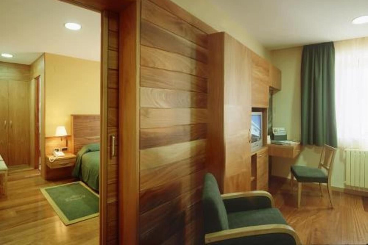 A hotel room with a wooden wardrobe, a bed with a green bedspread, a green armchair, a small table with a lamp, and a TV on a stand.