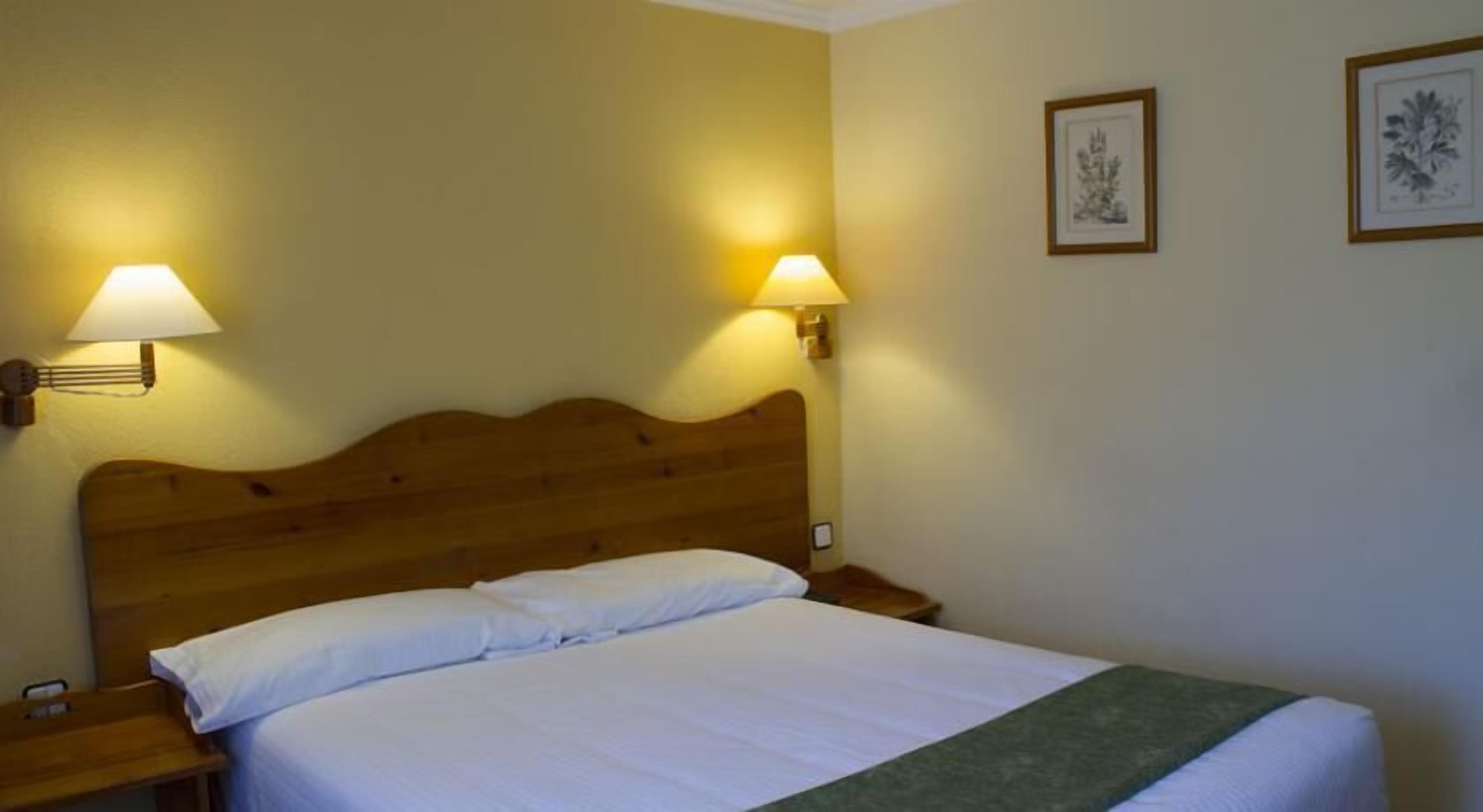 Habitación de hotel con cabecera de madera, dos lámparas de noche, cama con ropa blanca y dos cuadros en la pared.