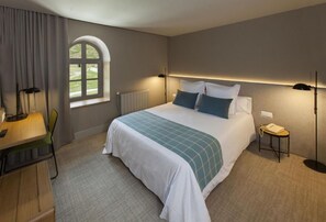 Habitación de hotel con cama, escritorio, silla, lámpara, mesita de noche y ventana.