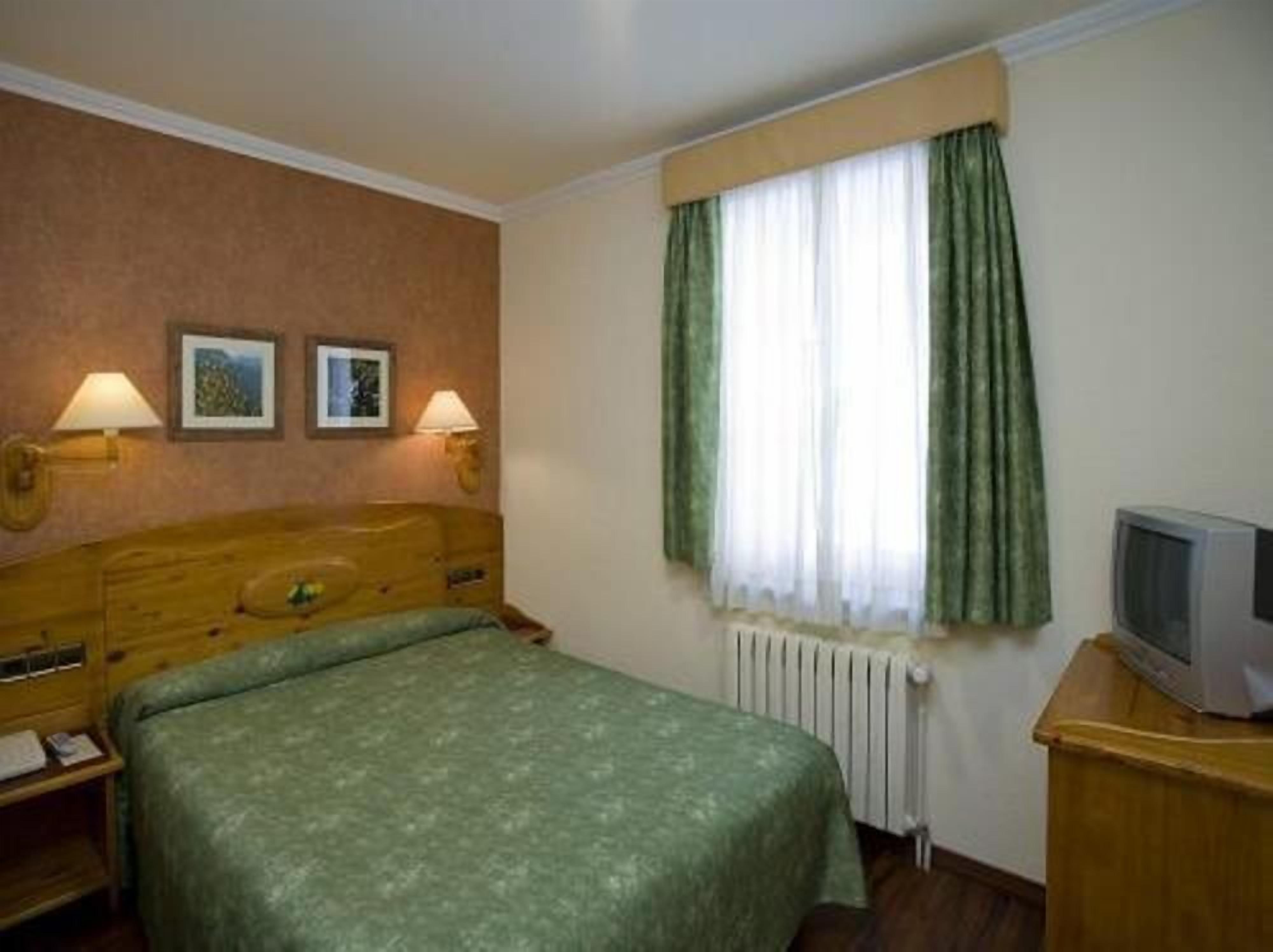 Una habitación de hotel con cama, dos lámparas de pared, televisor y ventana con cortinas.