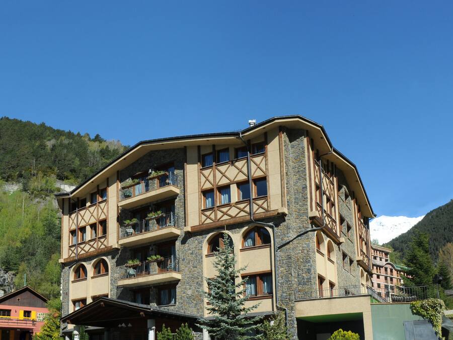Un hotel de varios pisos con fachada de piedra, balcones y montañas al fondo.