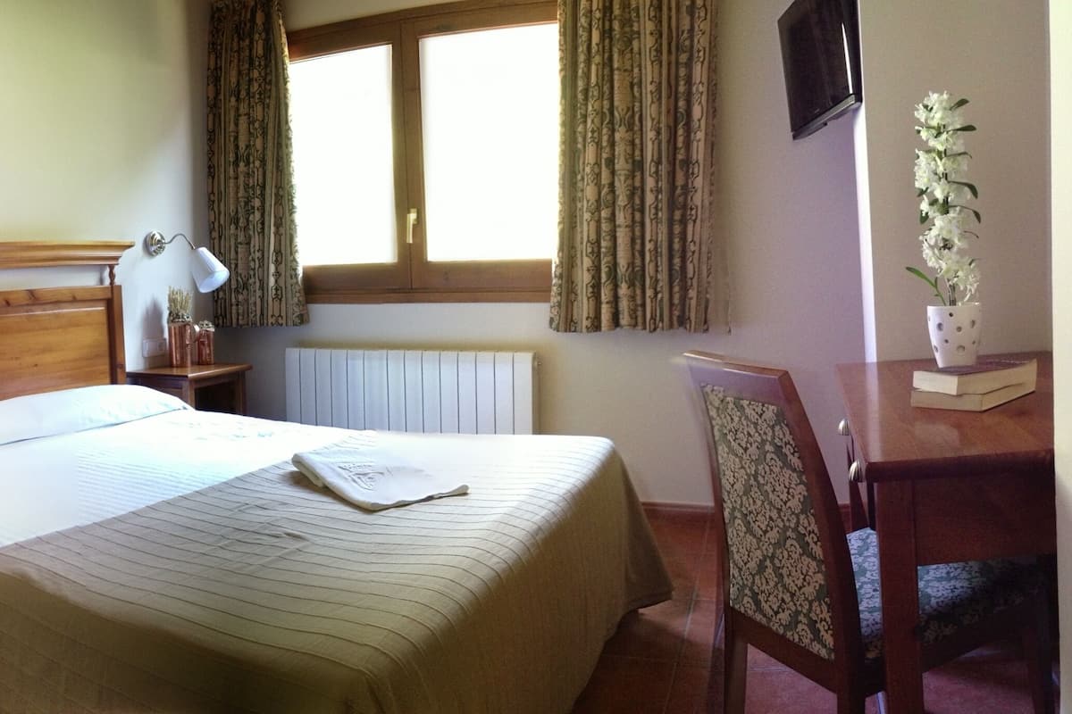 A neatly arranged hotel room with a bed, bedside table, lamp, chair, and a small table with a vase of flowers.