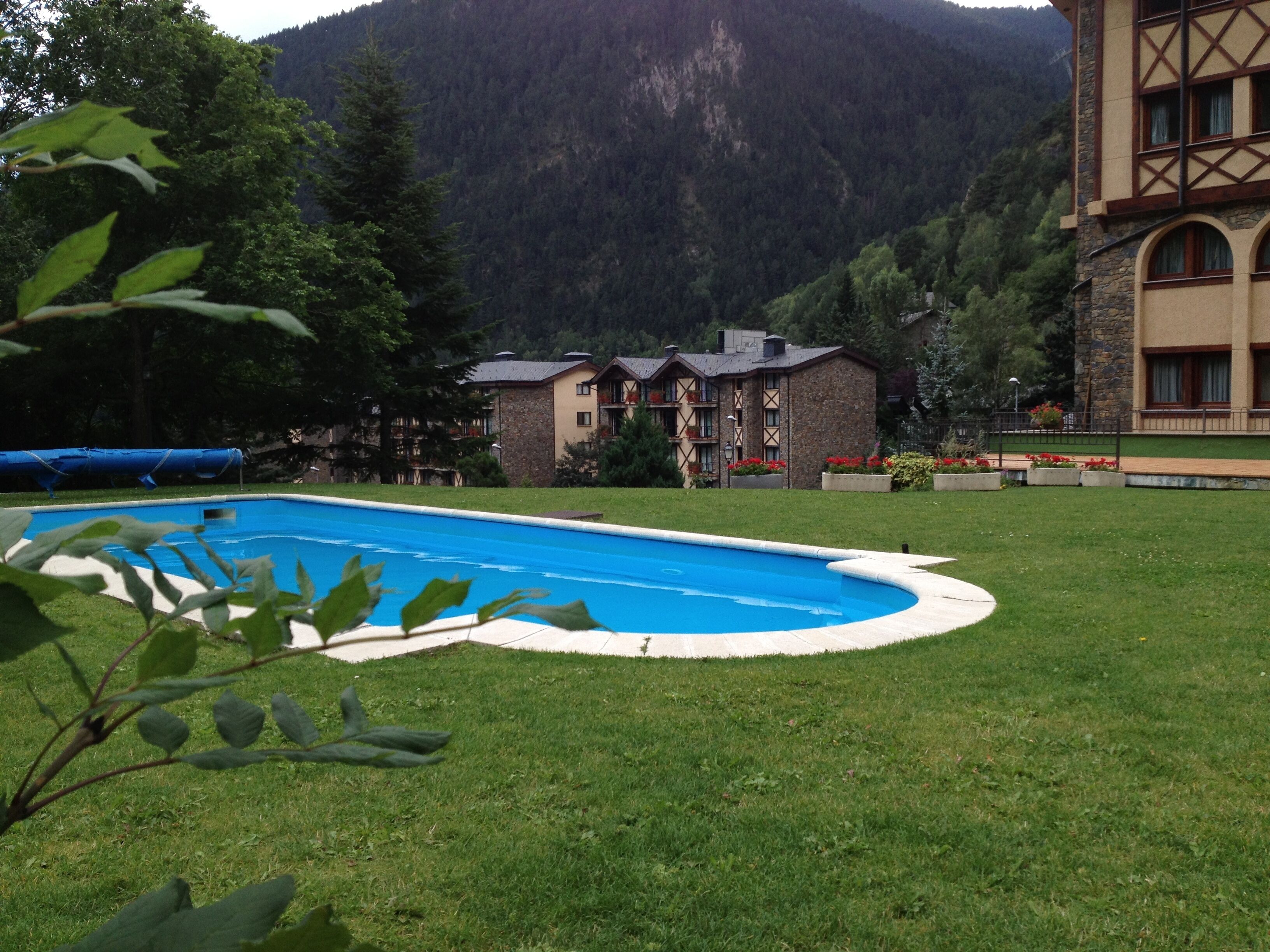 Una piscina rodeada de césped, con montañas al fondo y un edificio de fachada de piedra.