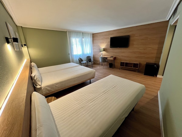 Habitación de hotel con dos camas individuales, un escritorio con una silla, un televisor montado en la pared y una pequeña zona de estar con sofá y mesa.