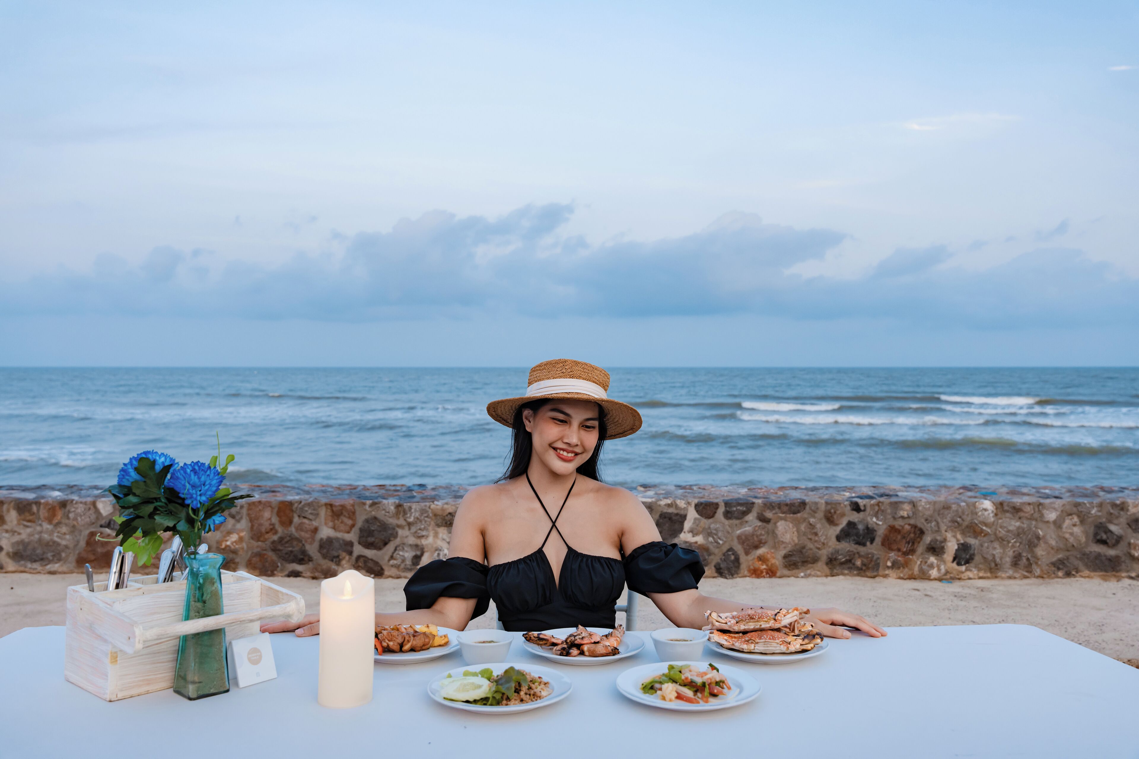A table set with plates of food, a candle, and a vase with flowers by the ocean.