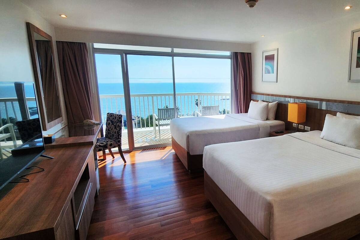 A hotel room with two beds, a desk with a laptop, a chair, and a balcony with a view of the sea.