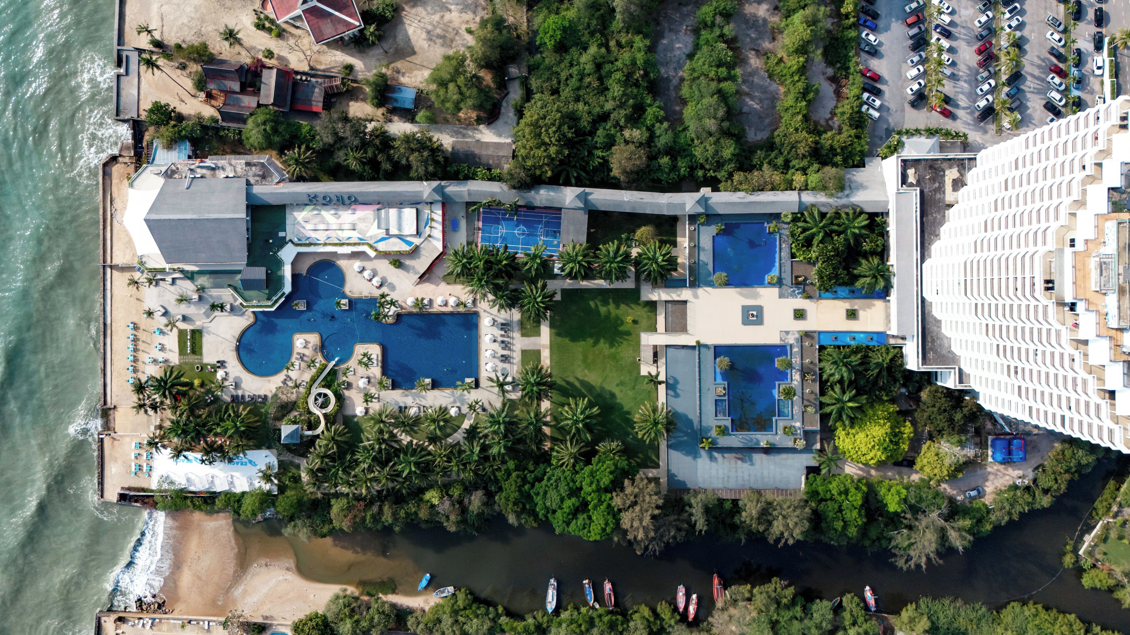 Eine Luftaufnahme eines Resorts mit mehreren Schwimmbecken, einem Strandbereich und einem Fluss mit Booten.