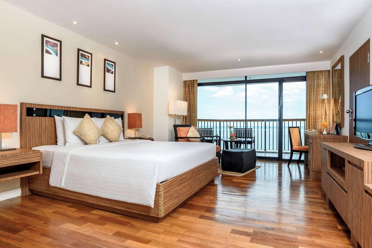 A hotel room with a large bed, a desk, a chair, and a TV. There is a balcony with a view of the ocean.