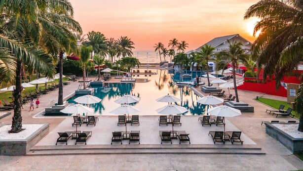 Un espace piscine aménagé dans un complexe hôtelier, avec de nombreux chaises longues et parasols, entouré de palmiers et offrant une vue sur le coucher du soleil.