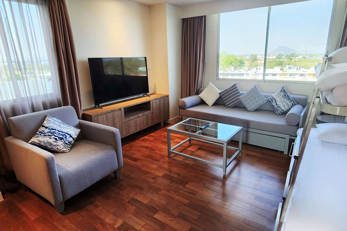 A hotel room with a sofa, a TV on a wooden stand, a bunk bed, and a window with a view of buildings and trees.