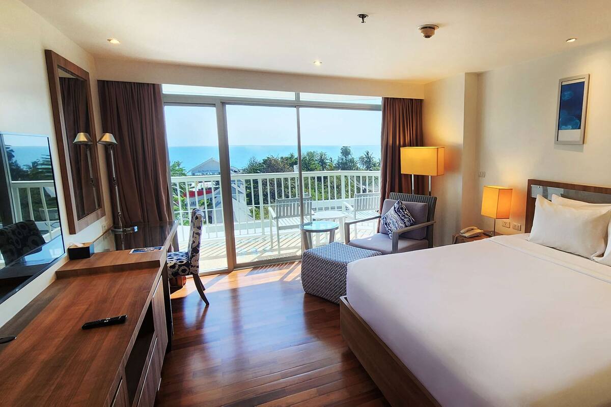 A hotel room with a large bed, a desk, a chair, and a balcony with a view of the ocean.