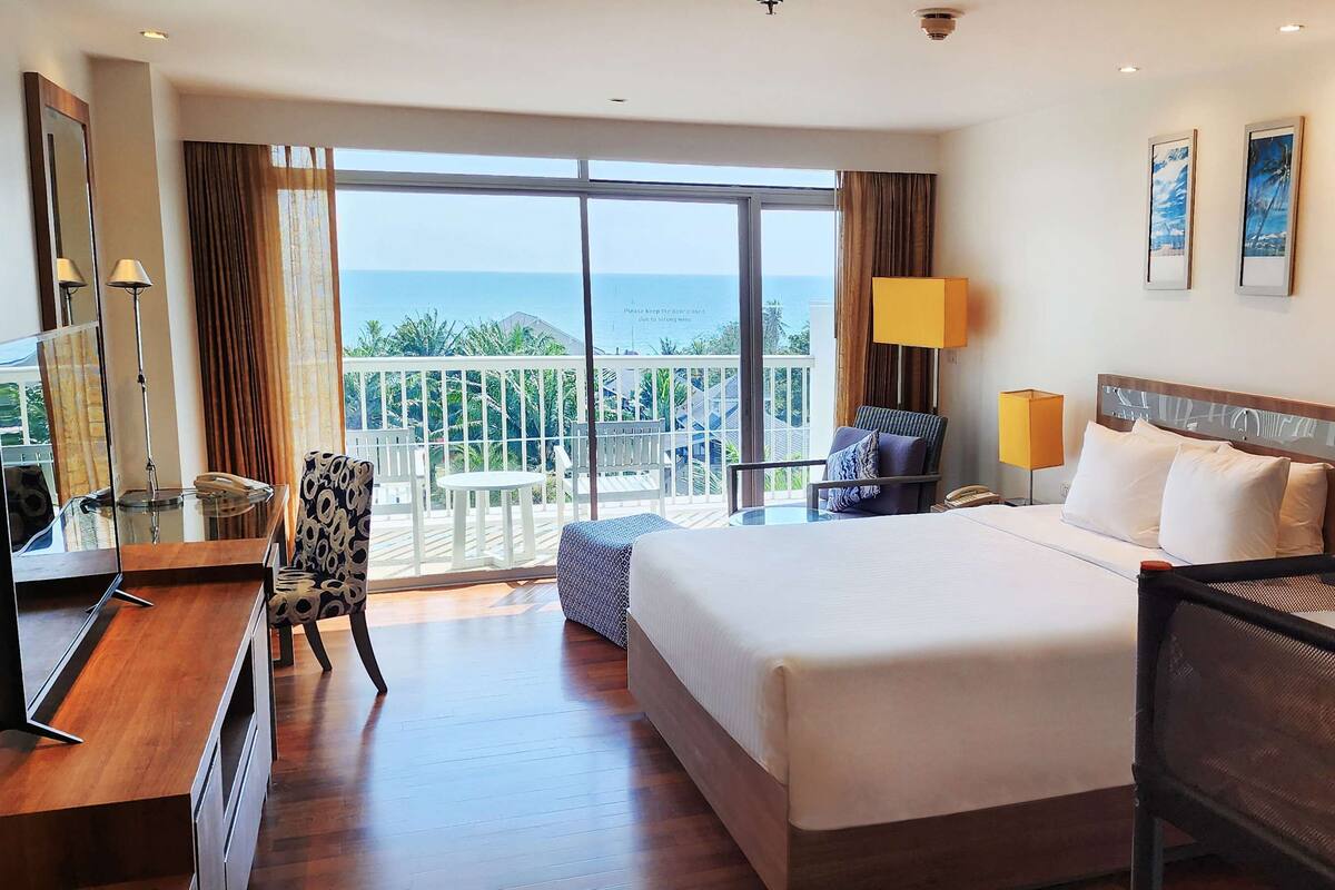 A hotel room with a large bed, a desk with a chair, a TV, and a balcony with a view of the sea.