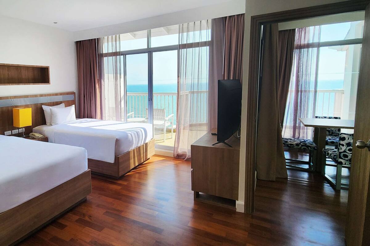 A hotel room with two beds, a TV, and a balcony with a view of the sea.