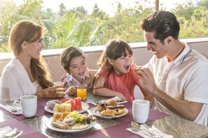 Une famille est assise à une table, en train de prendre son petit-déjeuner, et on aperçoit de la verdure à travers la fenêtre.