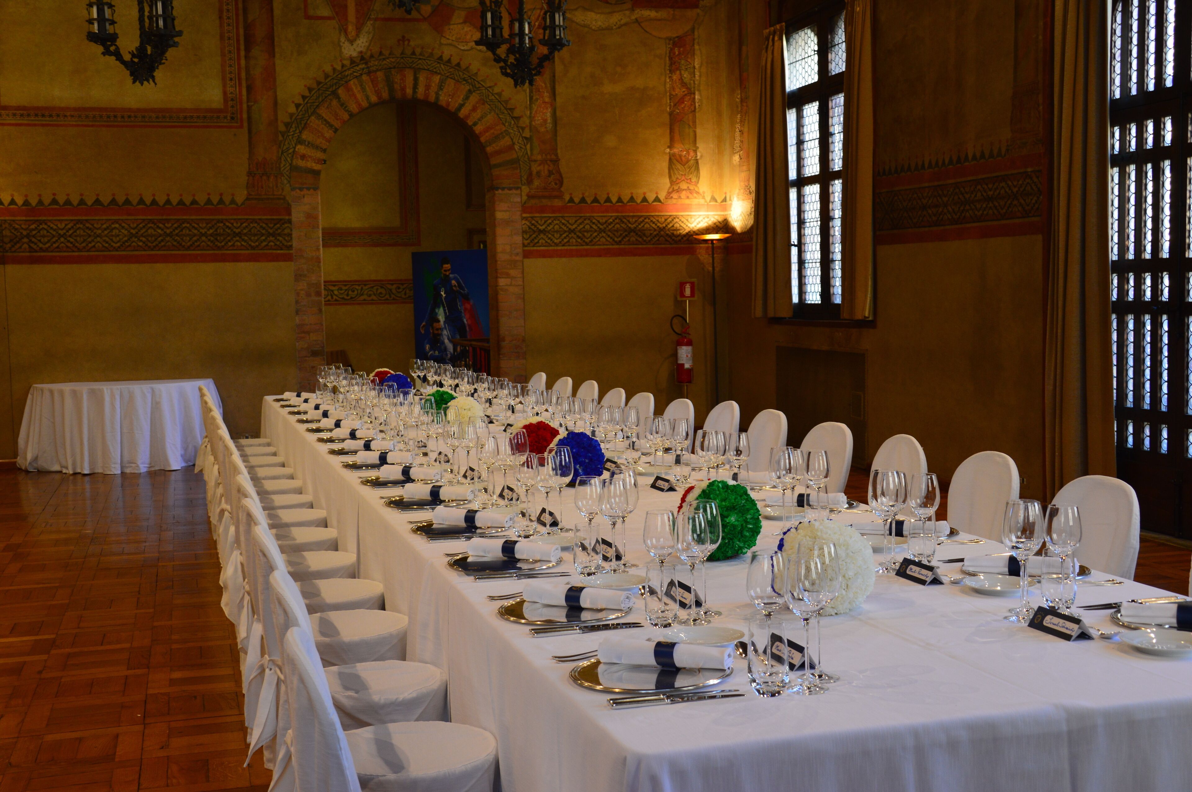 A banquet hall with a long table set for a formal event, featuring white tablecloths, chairs, and glassware.