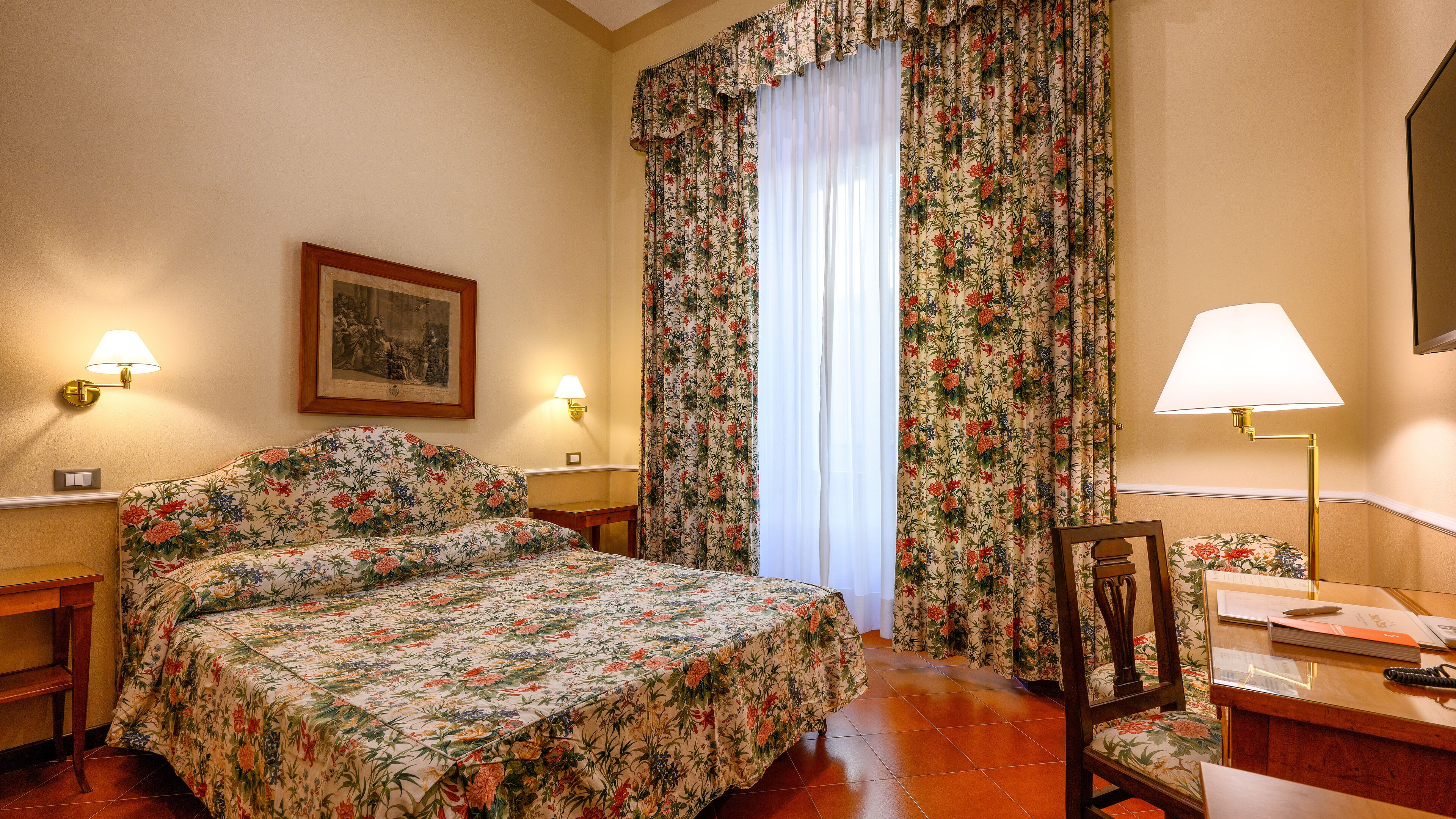 Una habitació d'hotel amb un llençol floral, un quadre al mur i una taula de nit amb una làmpada.