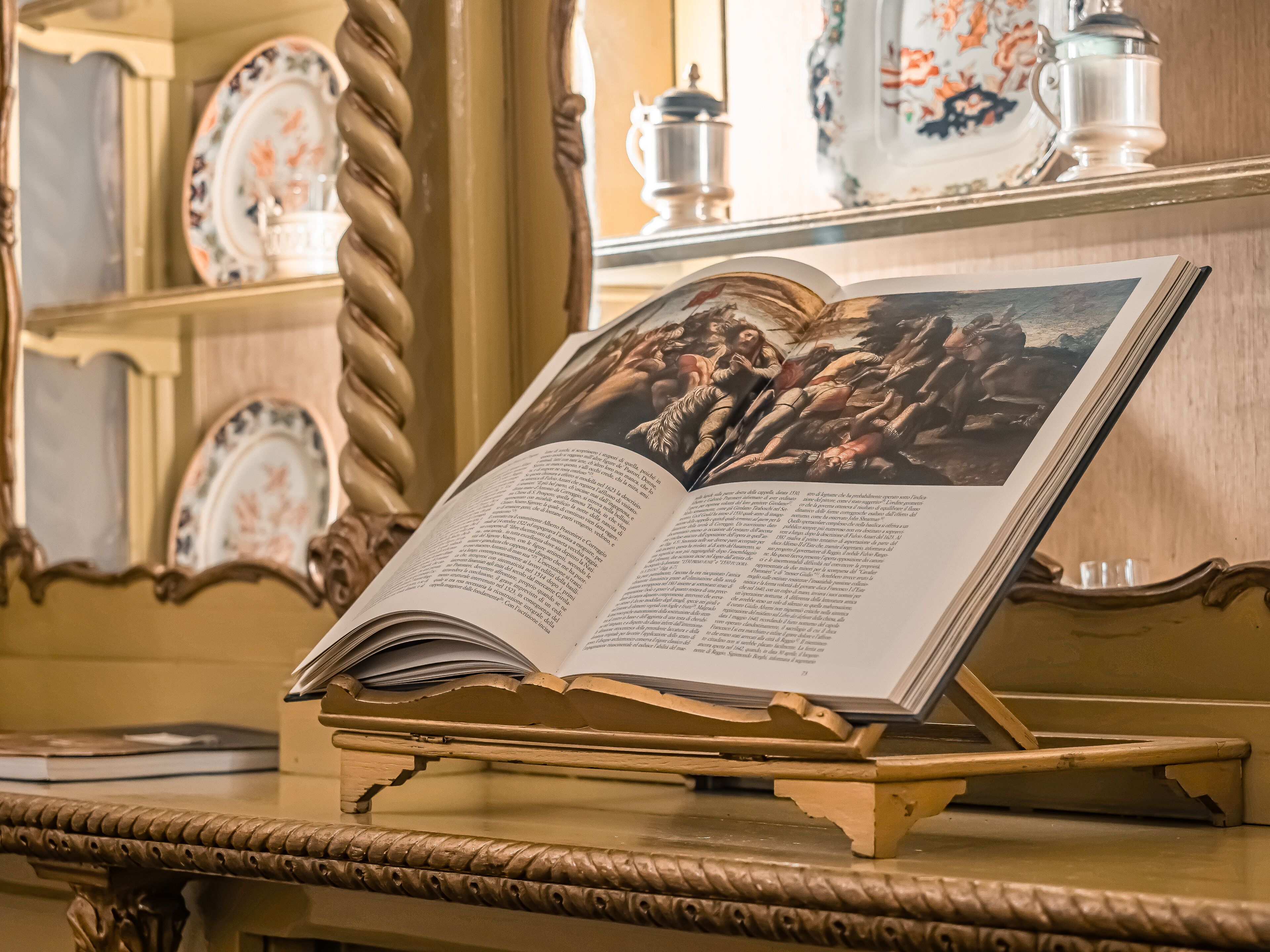 Een open boek op een standaard met decoratieve details, geplaatst op een tafel met een decoratieve spiegel en borden met een patroon erachter.
