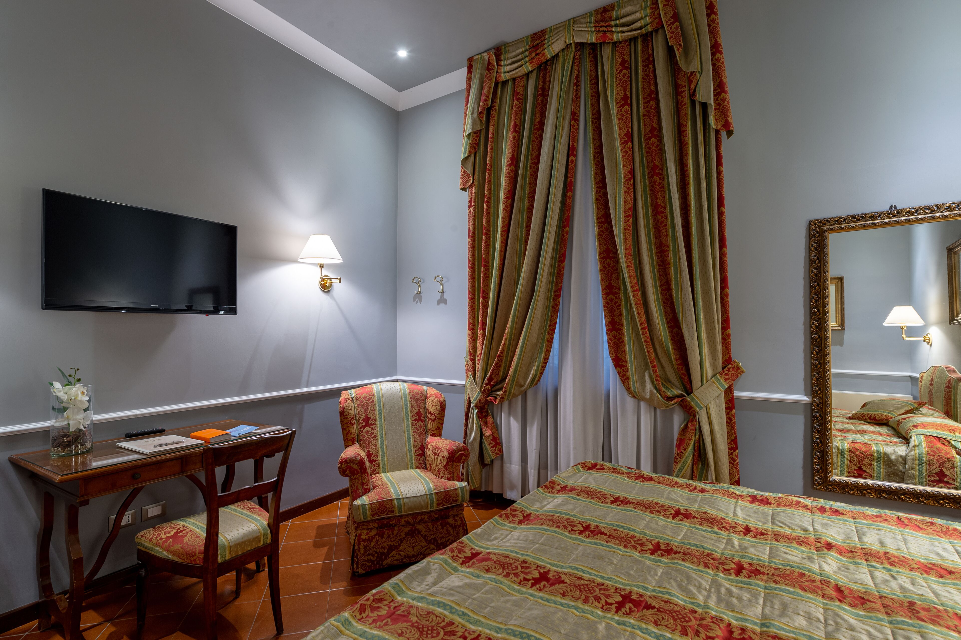 A bedroom with a bed, a desk with a chair, a TV mounted on the wall, and a large mirror.