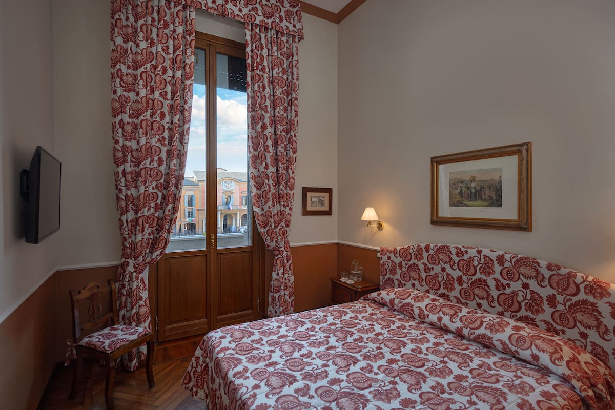 Un dormitorio con cama, una silla, una mesita de noche, una lámpara y un ventanal con cortinas.
