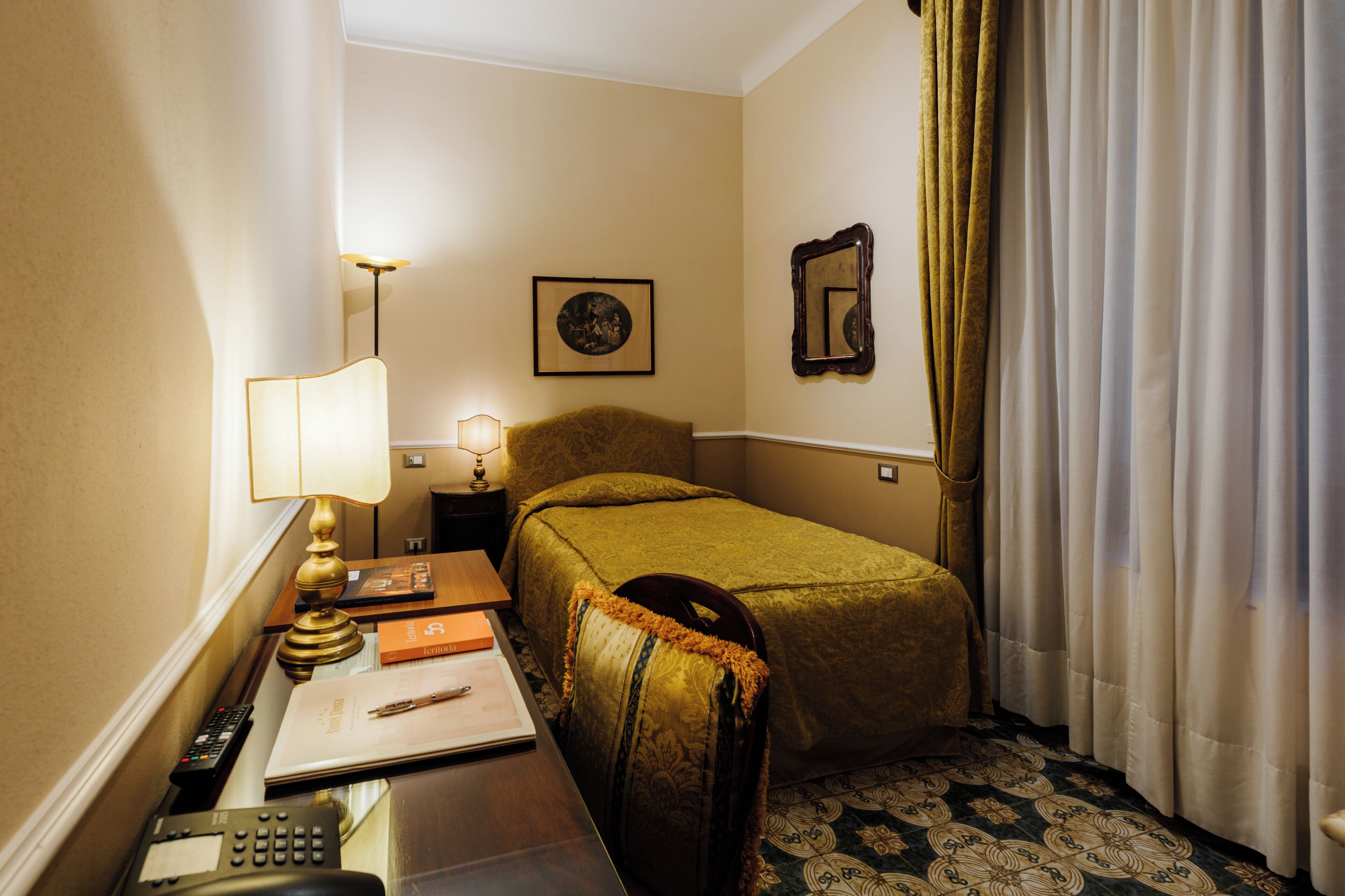 Ein Hotelzimmer mit einem Bett, einem Schreibtisch mit Lampe, einem Sessel, einem Telefon und einem gerahmten Bild an der Wand.
