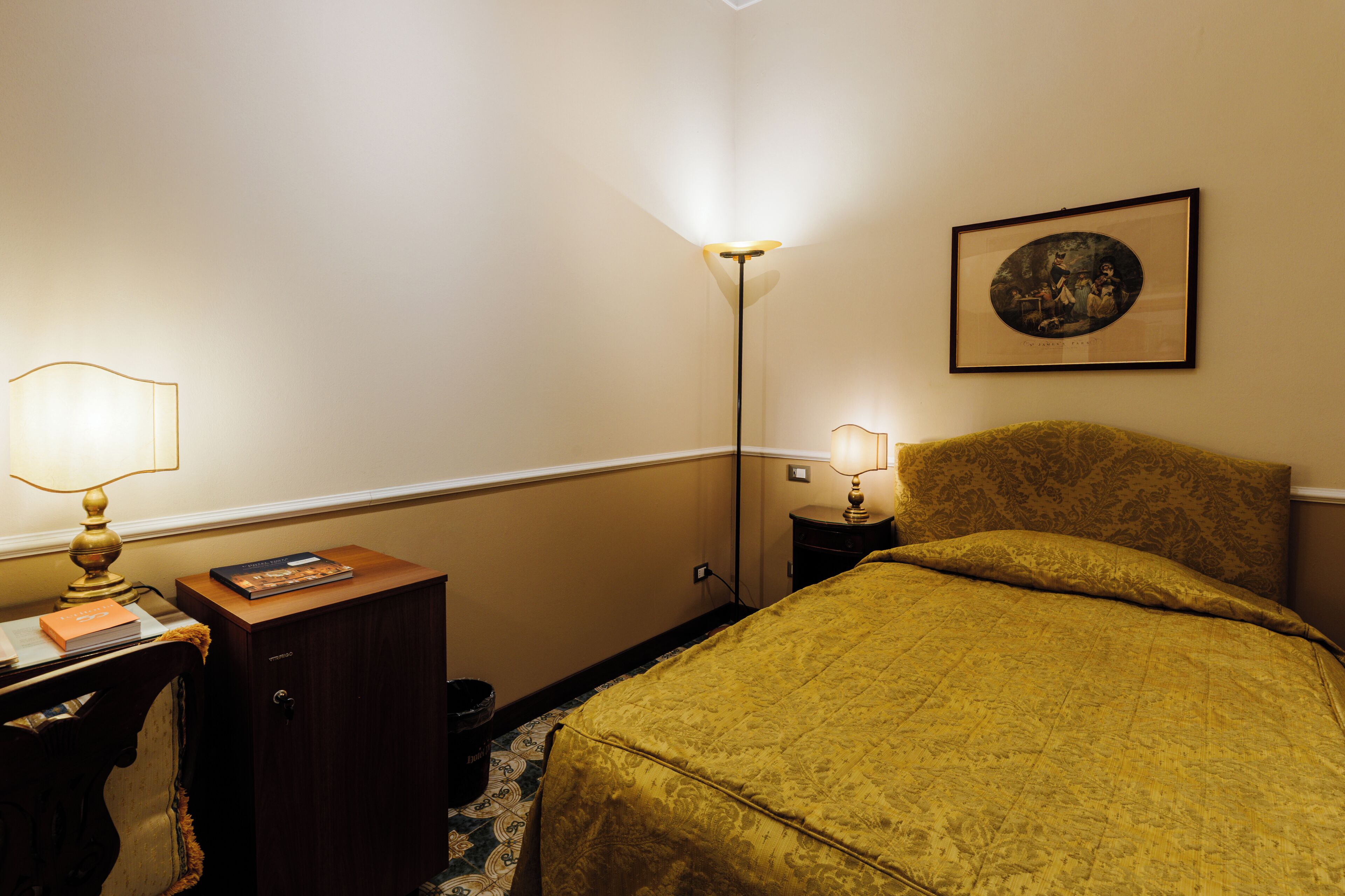 A hotel room with a bed, a desk with a lamp, a nightstand with a lamp, a framed picture on the wall, and a trash can.
