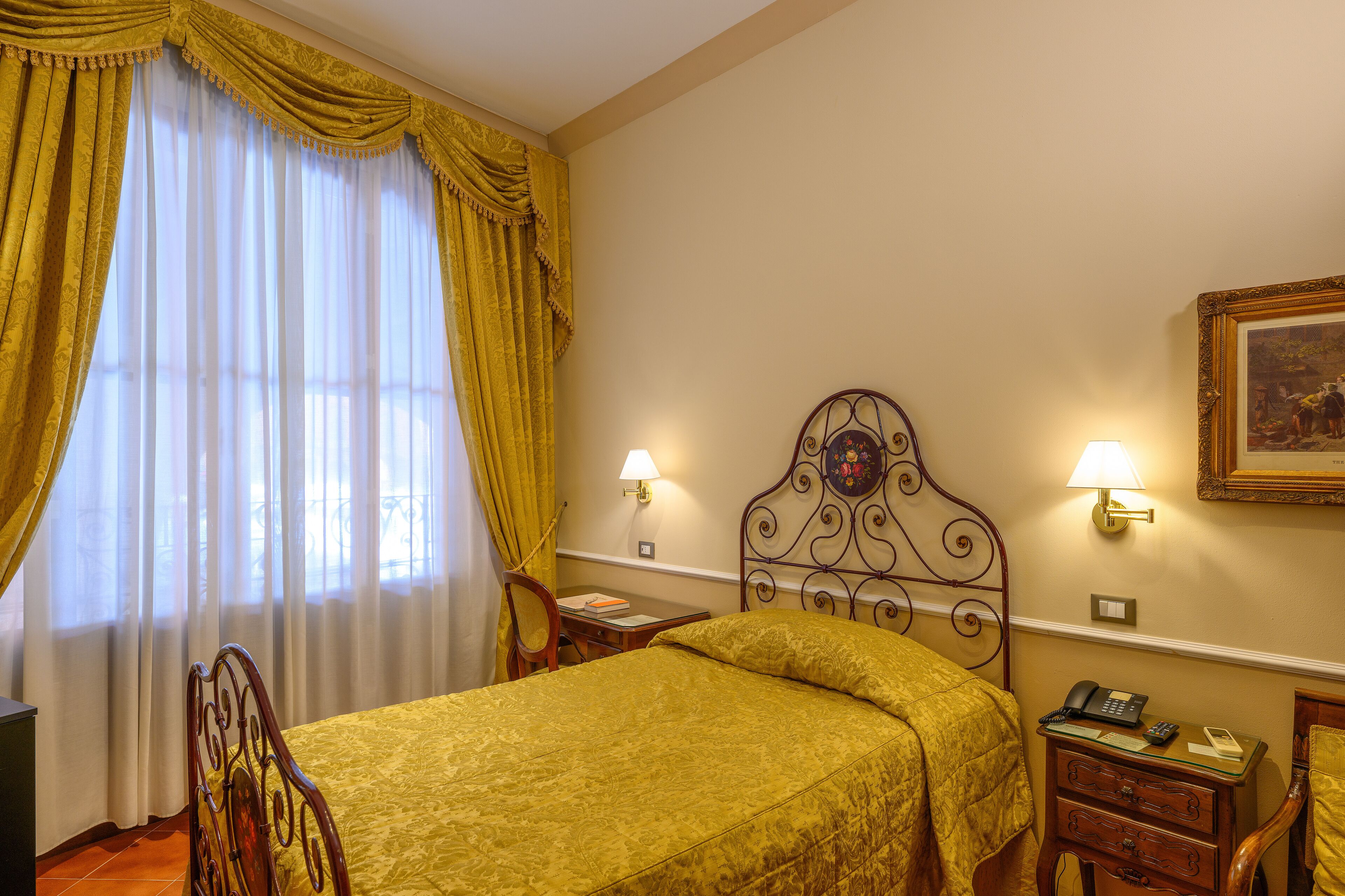 A hotel room with a bed, a desk, a chair, a nightstand, and a wall-mounted lamp.
