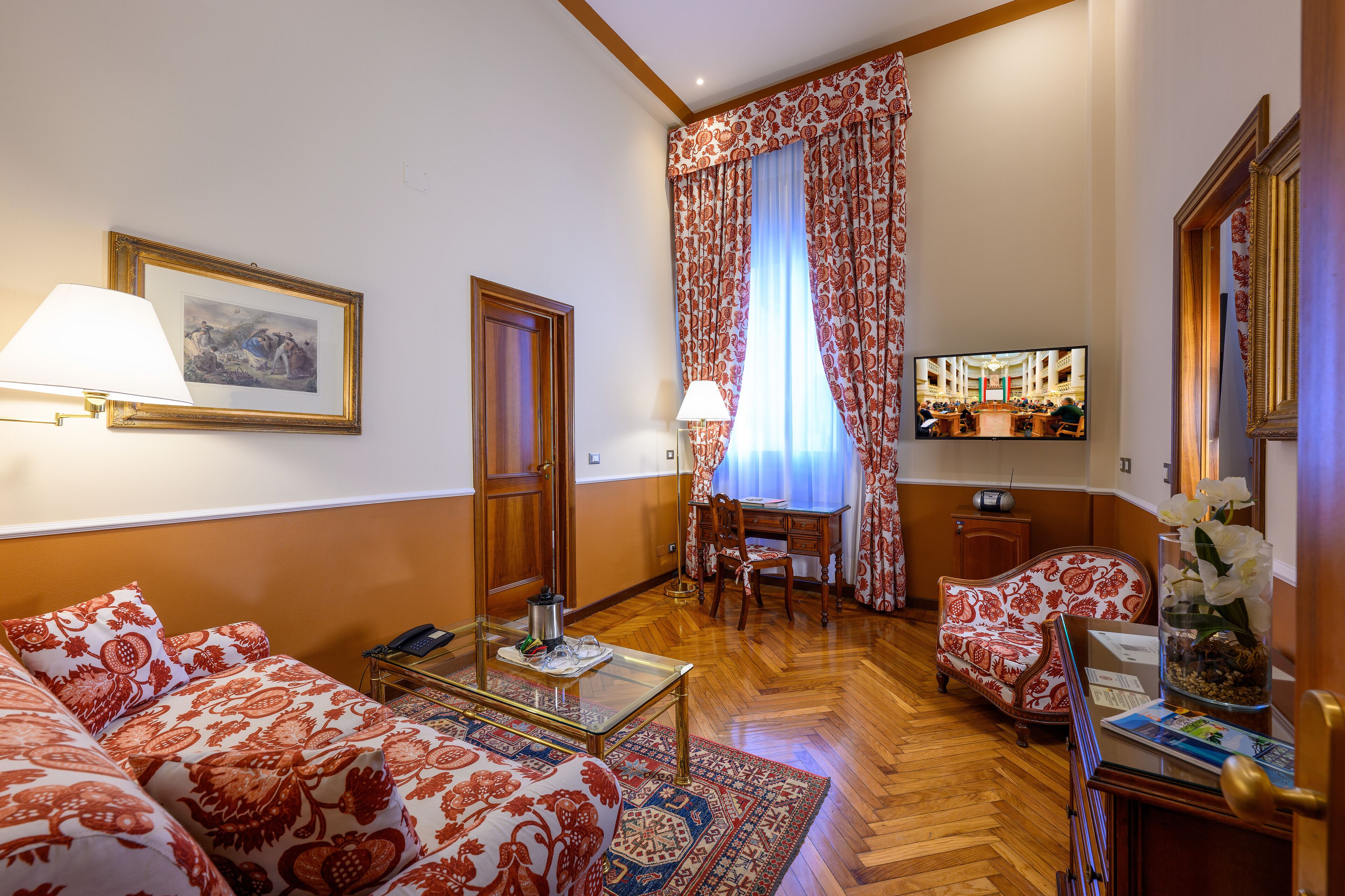 Habitación de hotel con sofá, una mesa de centro, escritorio y una silla. Hay un televisor en la pared y una ventana con cortinas.