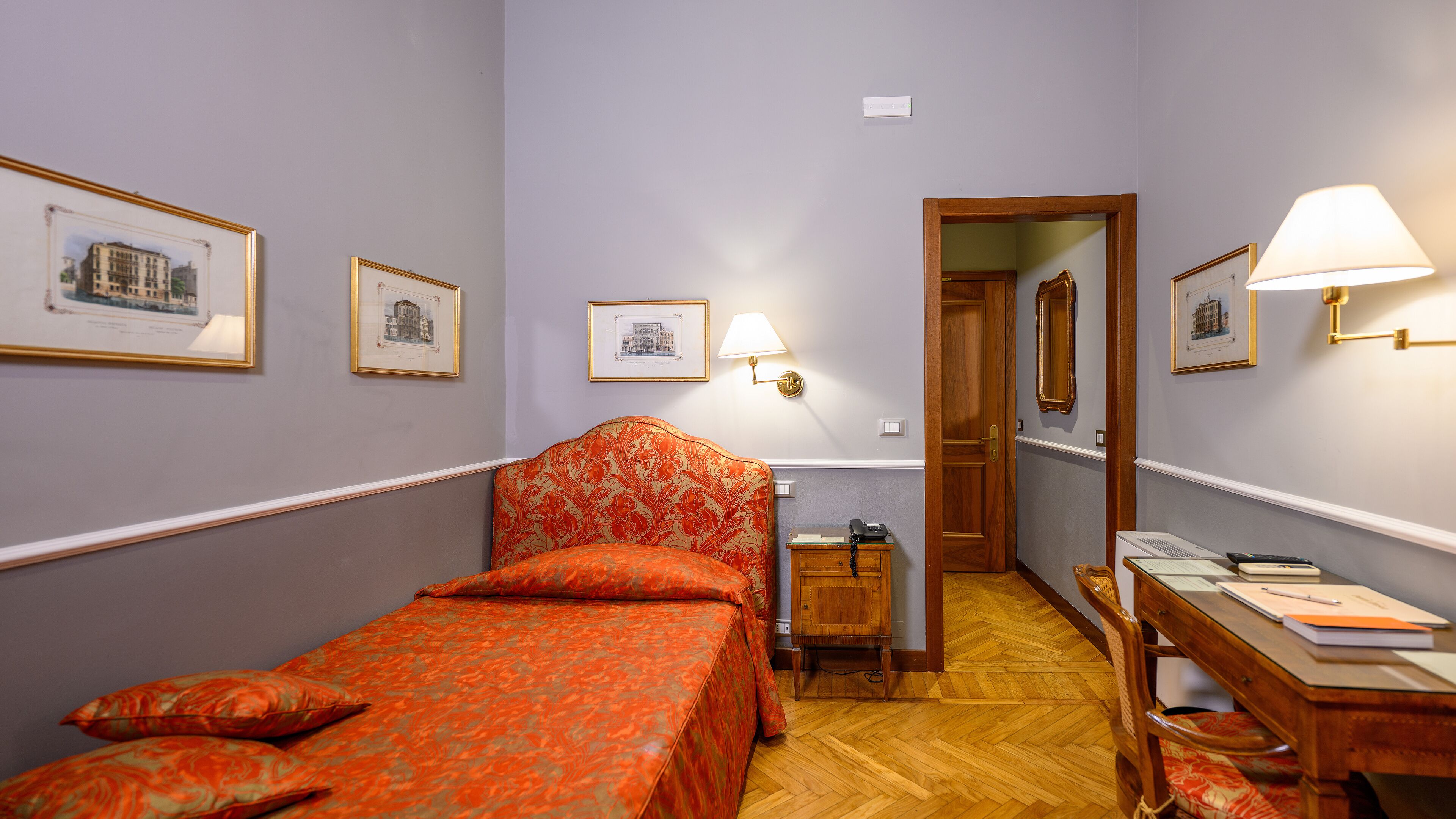 Habitación de hotel con una cama individual, un escritorio con silla, un armario de madera y cuadros enmarcados en las paredes.