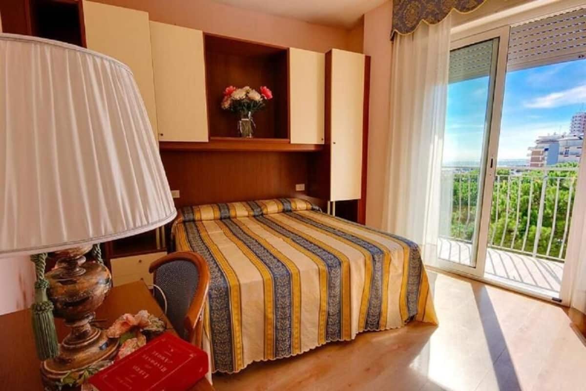 A bedroom with a bed, a desk with a lamp, a chair, a bookshelf, and a view of greenery through a balcony door.