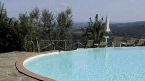 A swimming pool with a curved wooden edge, surrounded by stone tiles and a view of trees and hills in the background.