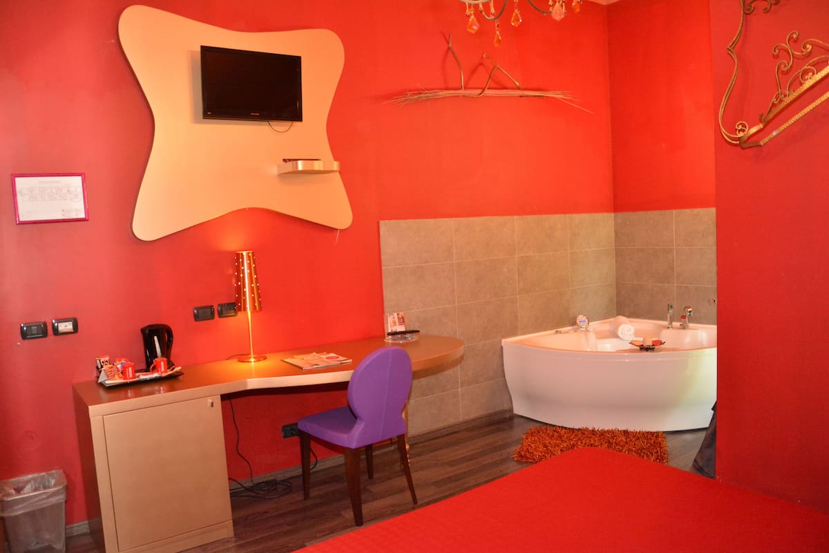 A modern bathroom with a red wall, a white bathtub, a wooden desk with a chair, and a small trash bin.
