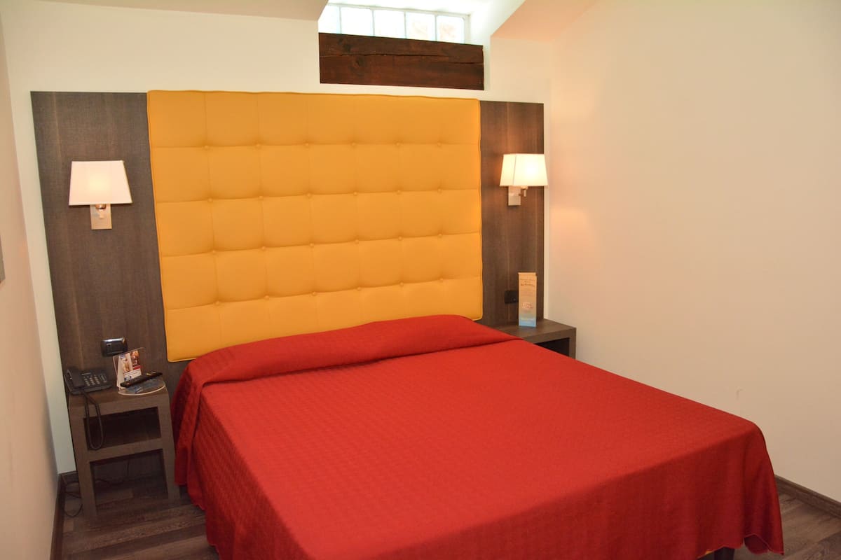 A hotel room with a large orange headboard, a red bedspread, two bedside lamps, a small table with a phone and a remote control, and a wooden headboard panel.
