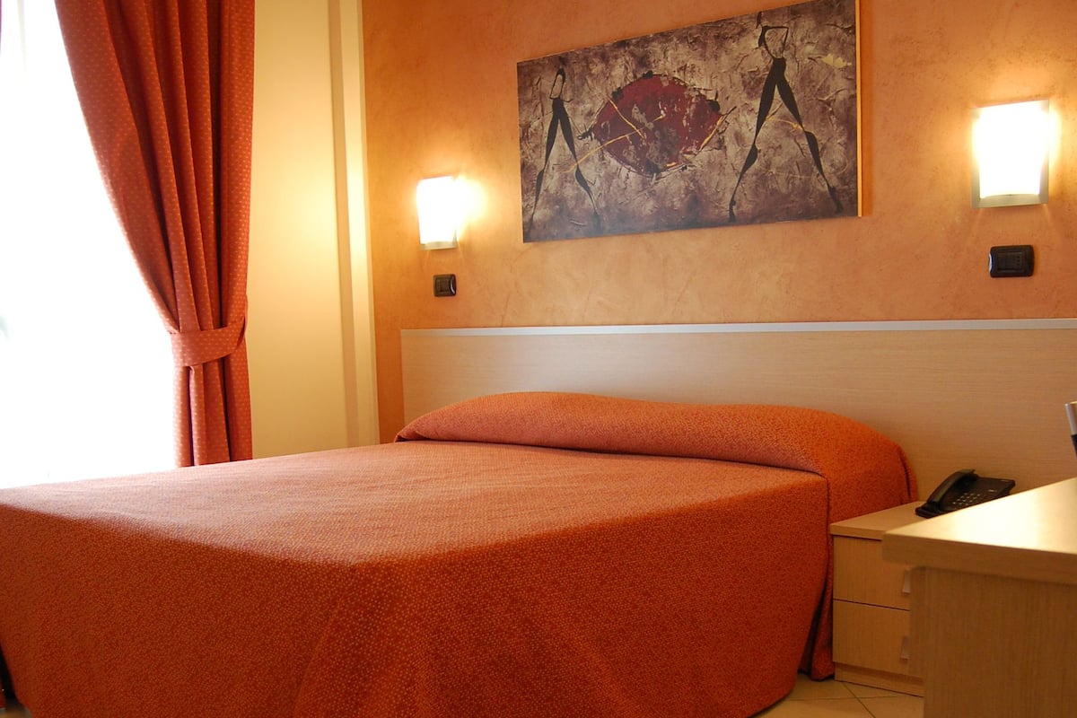 A hotel room with a bed, a desk, and a painting on the wall.