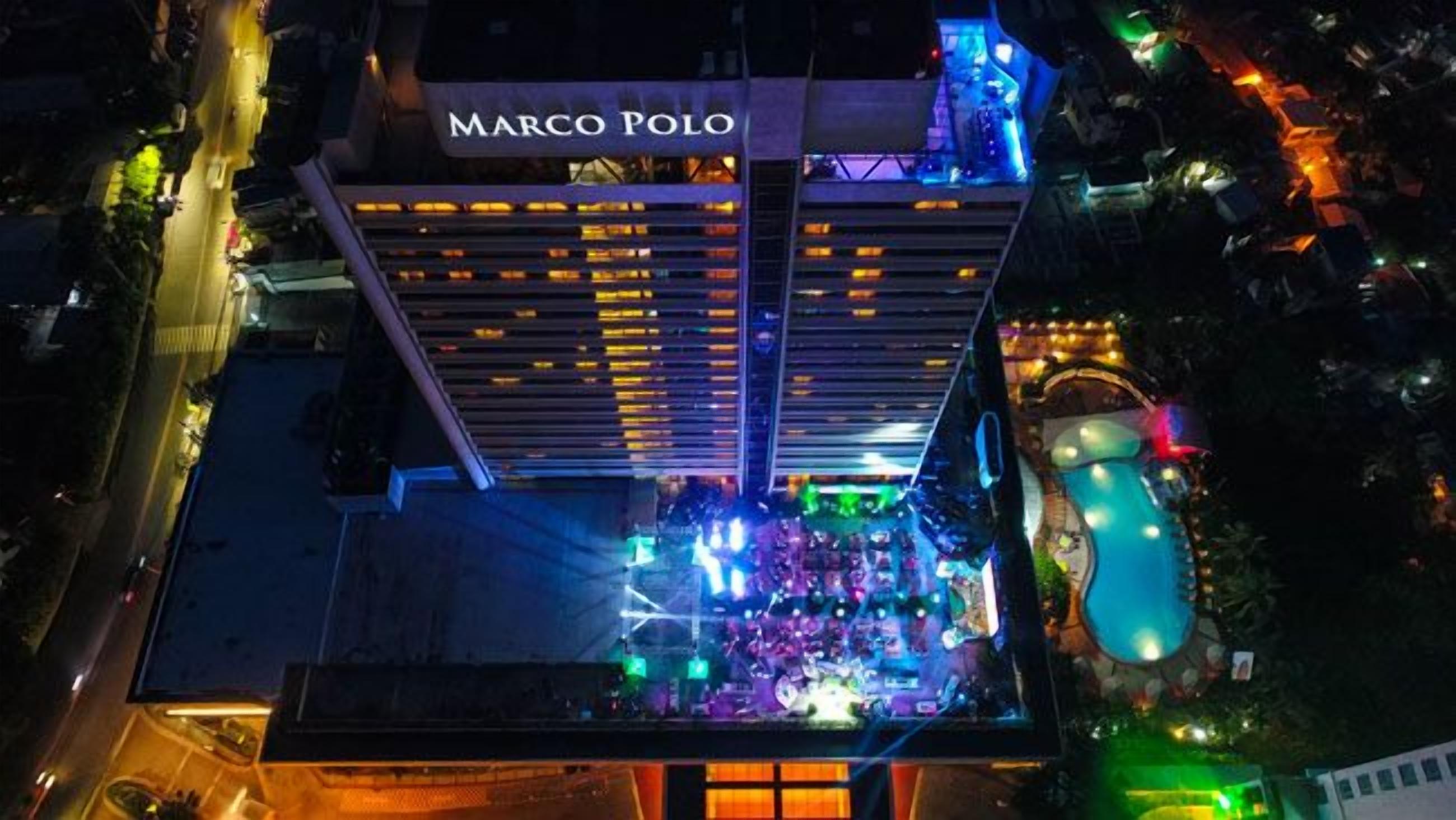 Un hôtel reconnaissable à son grand panneau « Marco Polo », doté d’une piscine sur le toit et d’une façade bien éclairée.