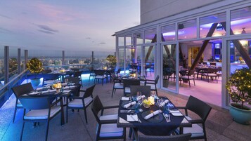 A rooftop restaurant with tables set for dining, overlooking a cityscape.